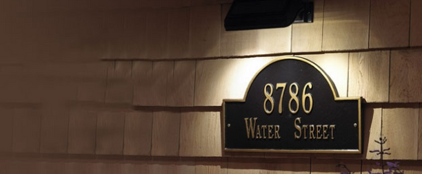 Arched black wall plaque with gold house number 8786 and the words Water Street, mounted on cream clapboard siding and lit from above