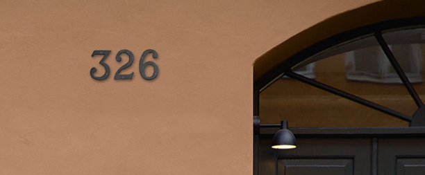 House number 326 in dark metal numerals on a terracotta stucco wall beside an arched doorway with a black gooseneck lamp